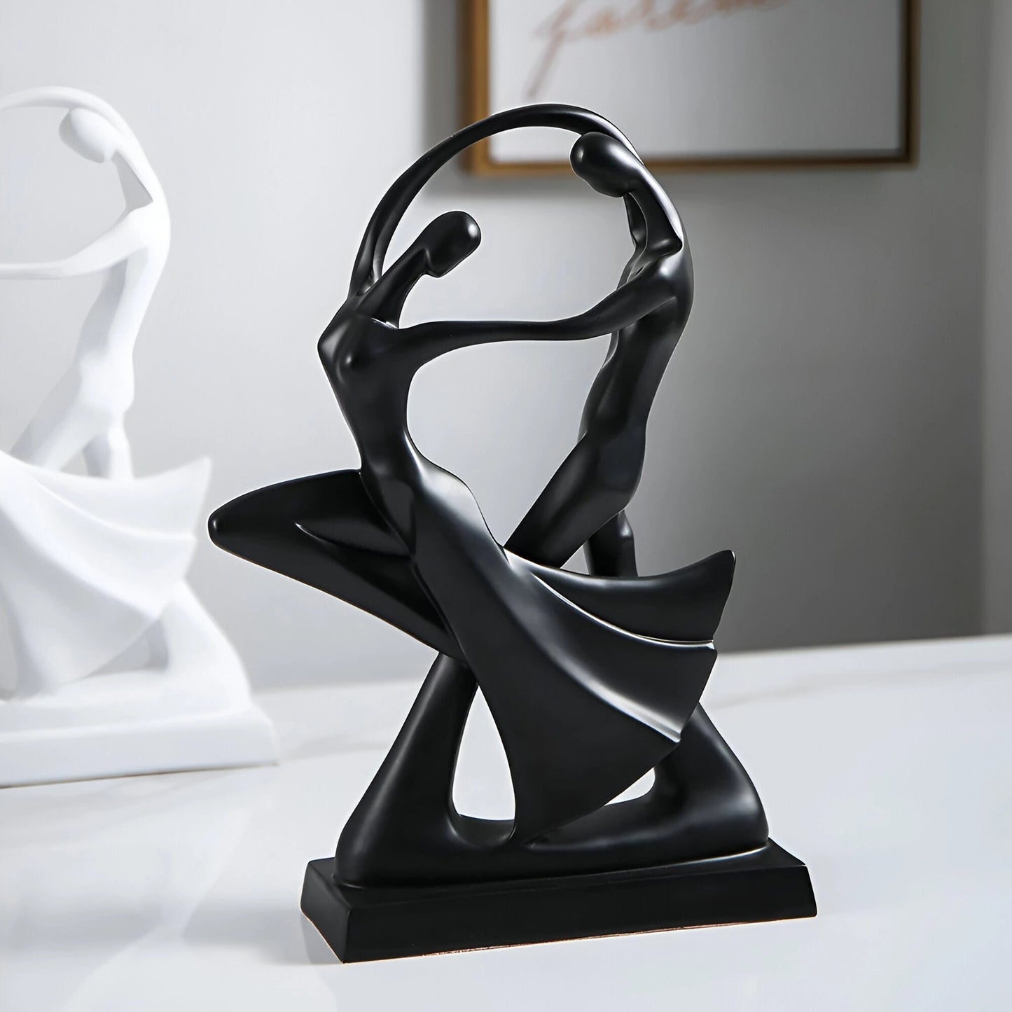 A modern black resin sculpture depicting an abstract couple in a graceful dance pose, measuring 30cm by 20cm. The artwork is designed with sleek, smooth lines and a metallic finish, presenting a contemporary piece ideal for art decor. The sculpture is displayed on a flat surface, highlighting its elegance and artistic expression.