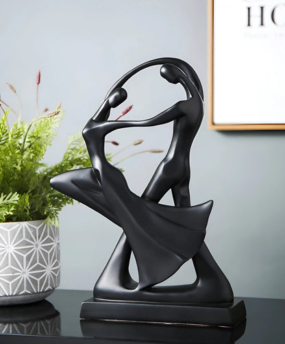 Abstract black resin sculpture of a modern dance couple, featuring sleek automotive design influences. The sculpture is showcased against a backdrop with plants and other decorative elements.